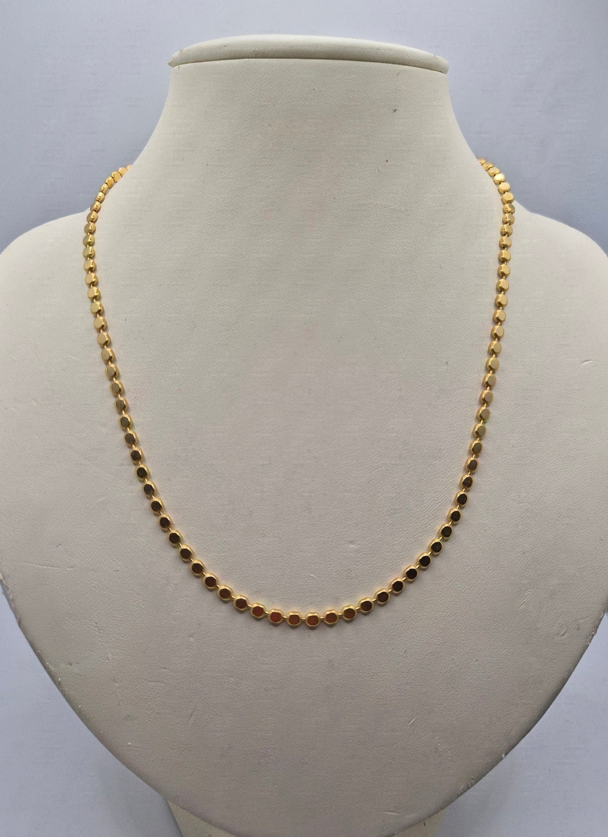 A gold beaded chain necklace displayed on a cream-colored jewelry bust against a light gray background.